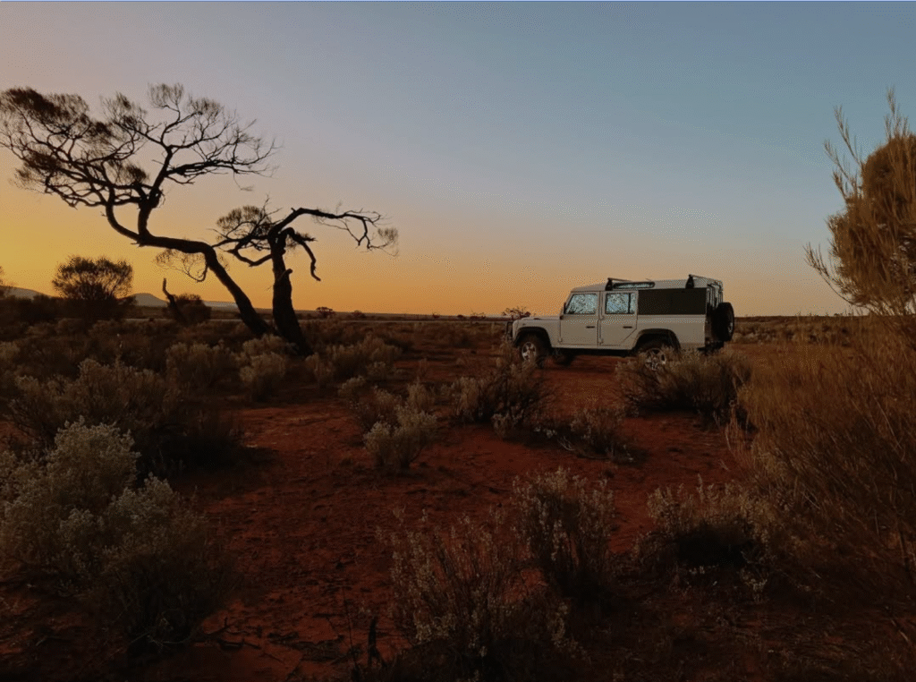 Land Rover Defender camper parked in the Australian outback at sunset with desert trees and red sand.